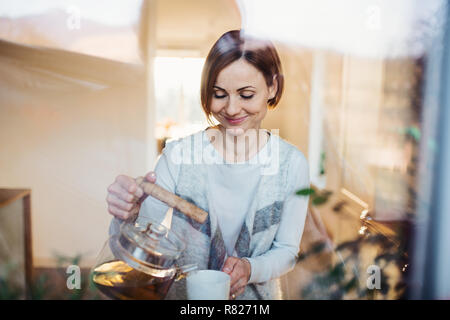 A young woman standing indoors in kitchen, pouring tea. Shot through glass. Stock Photo