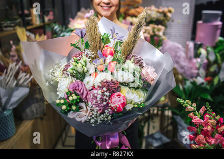 Nice and colorful bouqette of different flowers. Female florist hold it and smile. She is in flower shop. Cut view. Close up. Stock Photo