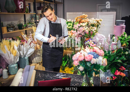 Picture of young male florist talk on phone and white down on black planchette. He has laptop, vase with flowers and paper rolls on table. He is busy. Stock Photo