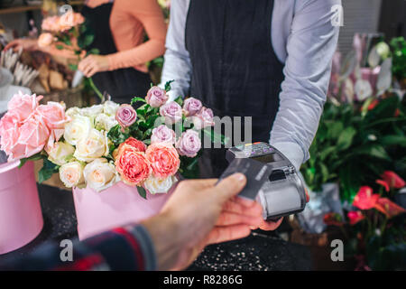 Cur view of man holding credit card in hand. He give it to florist. Flower seller has thereminal for cards. Table ull of colorful flowers. Stock Photo