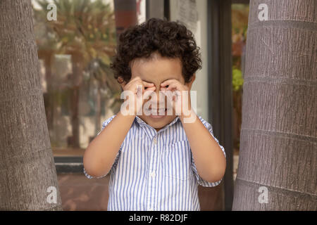A little boy pretends to use his hands as binoculars looking at me. Out on the street, a boy likes to use his imagination nestled between trees. Stock Photo