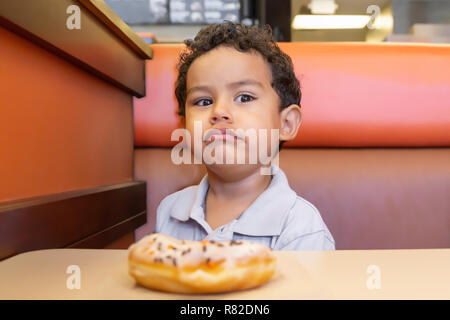 A little boy has a serious expression while he eats a donut in a booth. Head up, eyes focused to the side with a glaring stare. Stock Photo