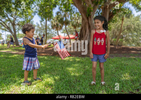 Hispanic brother and sister play at the community park under the shade of a large tree as he holds the American flag attached to a stick. Stock Photo