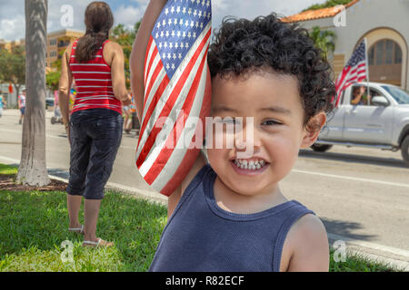 Fourth of July parade passes by as a boy toddler holds the American flag up looking at the camera with a big smile. Mother with her boy at the parade. Stock Photo
