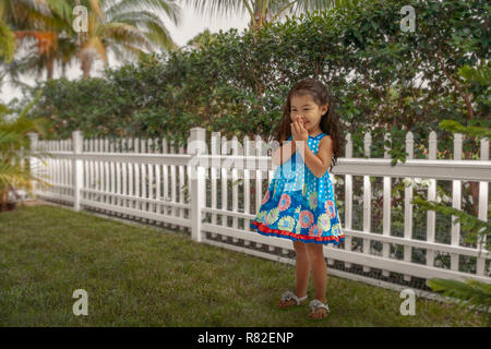 The child stands next to the white picket fence in her blue floral dress. She continues to smile looking at a distance cover her mouth with both hands Stock Photo