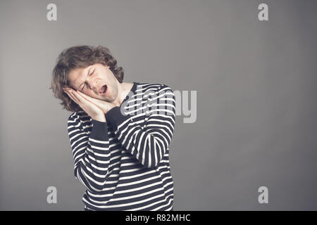 Young man in striped black and white sweater wants to sleep. Put his head in his hands and closed eyes while yawning. Standing on the grey background. Stock Photo