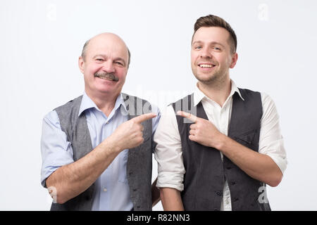 Two coworkers of different age pointing finger each other and looking at camera with happy face. Stock Photo