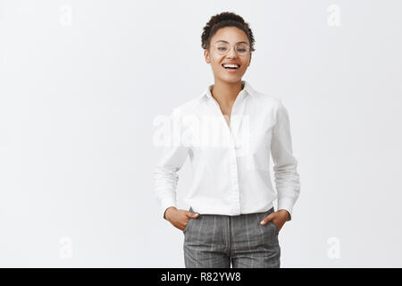 Everything under control. Good-looking african female in glasses, shirt and pants, holding hands in pockets, smiling and laughing with confident expression, triumphing, seeing great results of work Stock Photo