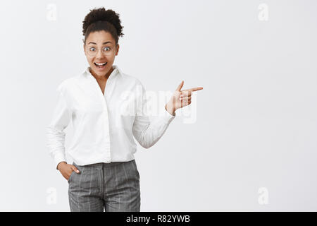 Cute dark-skinned female entrepreneur cannot believe she sees famous businesswoman during meeting, standing in trendy glasses and formal suit over gray background, pointing right with forefinger Stock Photo
