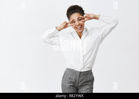 Charming happy girl living life to fullest. Cute dark-skinned female tutor in suit, showing peace signs over eyes, tilting head and smiling broadly, happy return home to kids after hard working day Stock Photo