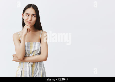 Hmm, let me think. Portrait of doubtful good-looking asian female in matching trendy outfit, smirking and gazing right while holding hand on chin, thinking, hesitating over gray background Stock Photo
