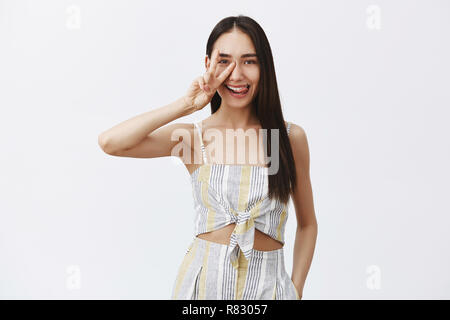 Indoor shot of emotive and playful cute female in elegant matching top with shorts, sticking out tongue flirty, smiling and showing peace sign over eye, having fun over gray background Stock Photo