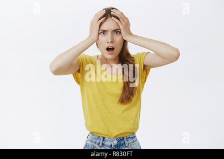 Girl having huge disaster on work feeling anxious and displeased holding hands on head in hopeless gesture opening mouth from surprise frowning standing dissatisfied and troubled over grey wall Stock Photo