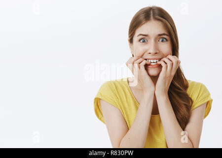 Waist-up shot of concerned anxious caucasian female employee scared of boss clenching teeth biting fingers from nerves popping eyes at camera feeling frightened and worried over gray wall, panicking Stock Photo