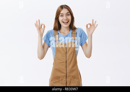 Everything okay, chill. Friendly and enthusiastic good-looking female in brown overalls showing ok gesture and smiling delighted giving positive feedback assuring she keeping things under control Stock Photo