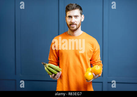 Colorful portrait of a caucasian bearded man in orange sweater holding fruits on the blue background Stock Photo