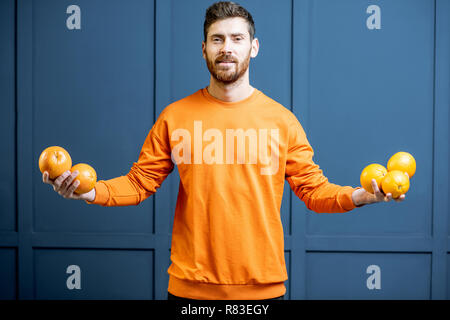 Colorful portrait of a caucasian bearded man in orange sweater holding fruits on the blue background Stock Photo