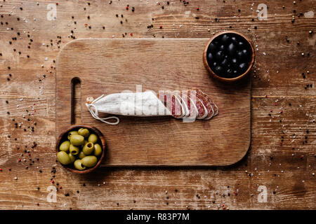 top view of cutting board with black and green olives in two bowls and delicious sliced salami on wooden table with scattered spices Stock Photo