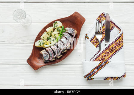 marinated herring, potatoes and onions in earthenware plate with glass of vodka, cutlery and embroidered towel on white wooden background Stock Photo