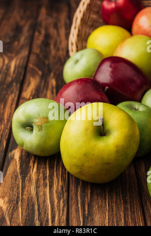 wicker basket with scattered yellow, green and red apples on wooden table Stock Photo