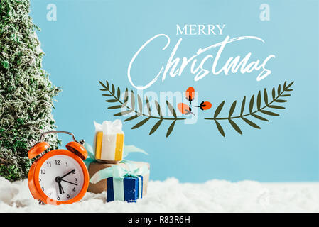 close-up shot of christmas gifts with alarm clock and miniature christmas tree standing on snow on blue background with 'merry christmas' lettering wi Stock Photo