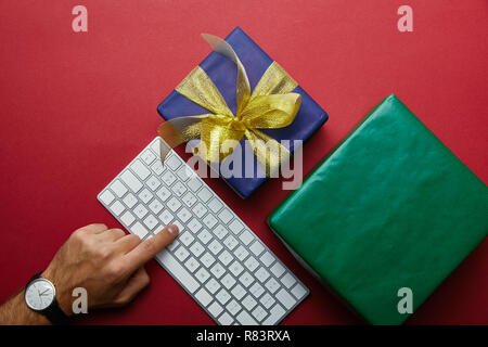 Top view of man pushing button on white computer keyboard near colourful gifts on red background Stock Photo