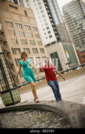 Young man holding his girlfriend's hand as she walks along the wall of a fountain. Stock Photo