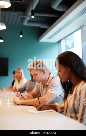 Group Of Young Candidates Sitting At Boardroom Table Making Notes At Presentation At Business Graduate Recruitment Assessment Day Stock Photo