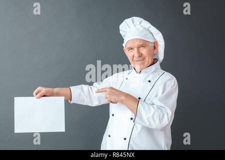 Senior chef studio standing isolated on gray pointing at paper looking camera playful Stock Photo