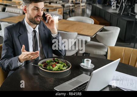 Businessperson on business lunch at restaurant sitting eating salad drinking espresso discussing work on smartphone Stock Photo