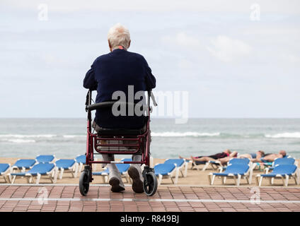 Elderly man sitting on wheel walker/rollator looking out over beach as people sunbathe. Stock Photo