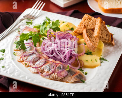 Appetizer of salted herring, red onion, potatoes and rye bread in white plate on dark brown wooden table in restaurant Stock Photo