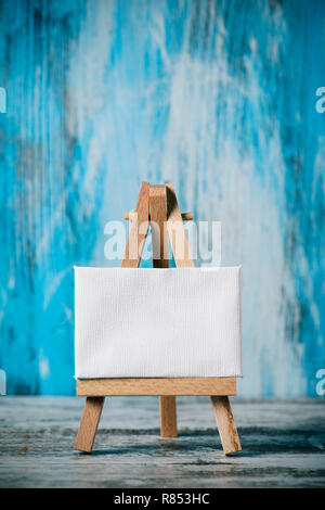 a blank white canvas on a wooden easel, on a wooden surface, against a blue rustic wooden background Stock Photo