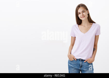 Charming shy and cute european young female brunette in casual blouse standing with hands in pockets over white background tilting head and smiling with friendly look getting know new coworkers Stock Photo