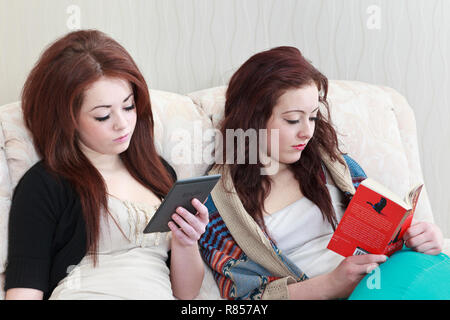 Two teenage twin girls aged 15 years one reading a Kindle and the other reading a paperback book whilst both sitting on a sofa together. UK Britain Stock Photo