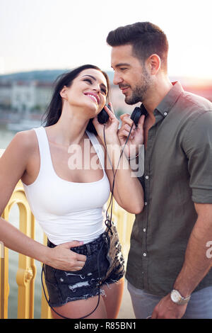 Happy young trendy caucasian couple sharing headphones at outdoors, listening to music Stock Photo