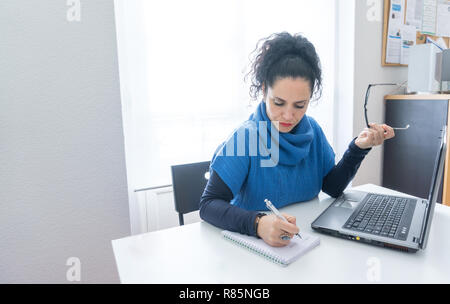 Middle age woman with black hair and white skin working on papers paperwork and the laptop in an office with red lips holding her glasses Stock Photo