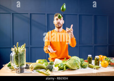 Conceptual portrait of a vegetarian man in bright sweater throwing up vegetables on the blue background Stock Photo