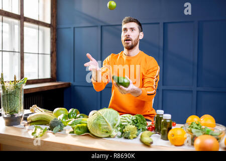 Conceptual portrait of a vegetarian man in bright sweater throwing up vegetables on the blue background Stock Photo