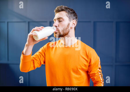 Conceptual portrait of a sports man in bright sweater drinking milk from the bottle on the blue background Stock Photo