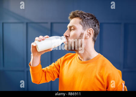 Conceptual portrait of a sports man in bright sweater drinking milk from the bottle on the blue background Stock Photo