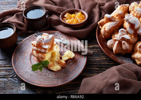 Profiterole with custard cream filling served for breakfast on a earthenware plate on a dark wooden table with brown cloth, apricot jam and mugs with  Stock Photo