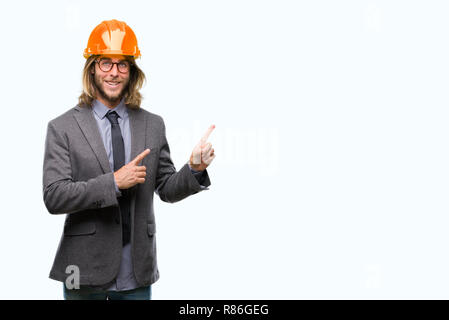 Young handsome architec man with long hair wearing safety helmet over isolated background smiling and looking at the camera pointing with two hands an Stock Photo