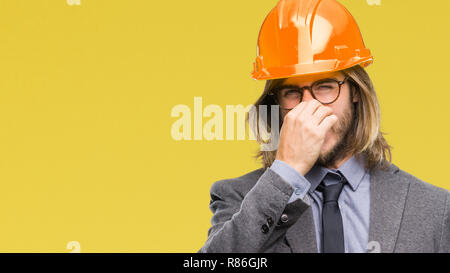 Young handsome architec man with long hair wearing safety helmet over isolated background smelling something stinky and disgusting, intolerable smell, Stock Photo