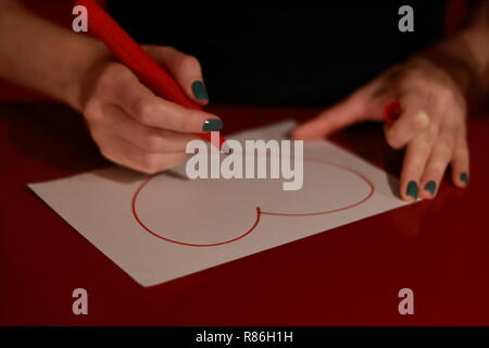 Girl drawing a heart on a white paper with a red marker. Hands with green nail and marker. White paper on a table with drawing. Stock Photo