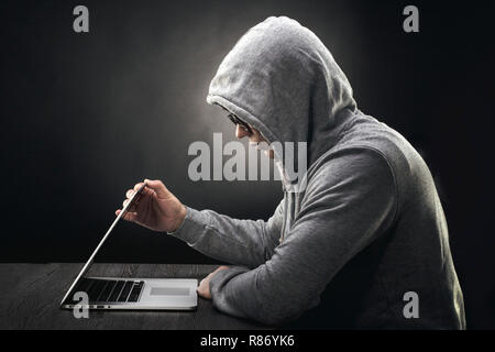 a hacker in a hood in profile sits at a laptop and starts to close it Stock Photo