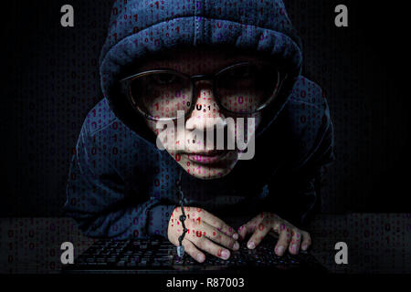 binary code on the background of a hacker in a hood and glasses sits at the keyboard and looks strangely into the camera Stock Photo