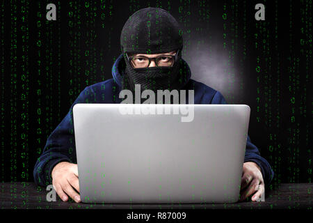 binary code on the background of a cybercriminal in a balaclava and glasses sits at a laptop Stock Photo