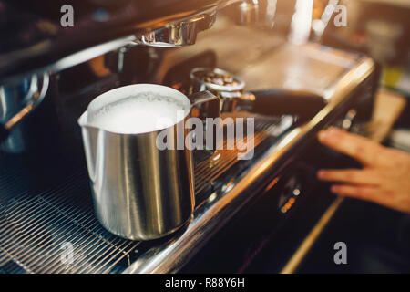 Close up of metal cup with hot milk in it. It stands on coffee machine at cezve. Guy touches machine Stock Photo