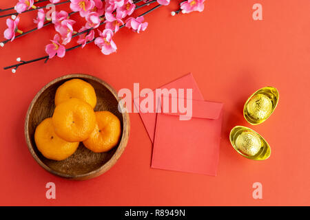Flat lay Chinese New Year Background - Cherry Blossom, Mandarin Orange, Red Envelop and Golden Ingots on red background. Stock Photo